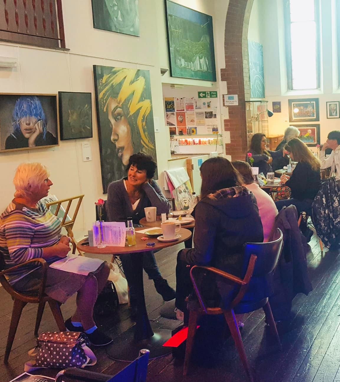 The cafe area of Barnabas Arts House, visitors at round wooden tables under three large portrait paintings, including a yellow-haired figure on the centre wall.