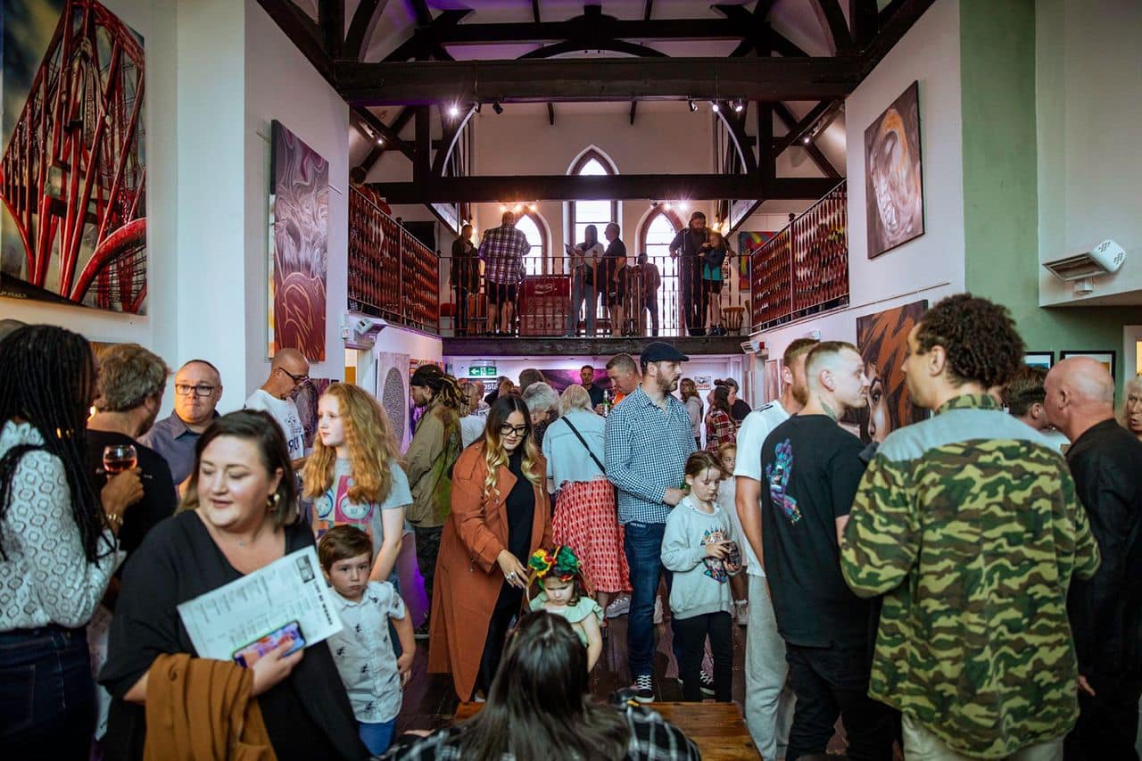 Opening night at Barnabas Arts House. Visitors fill the main nave floor and the gothic mezzanine balcony, exposed roof trusses overhead, large paintings on the walls.