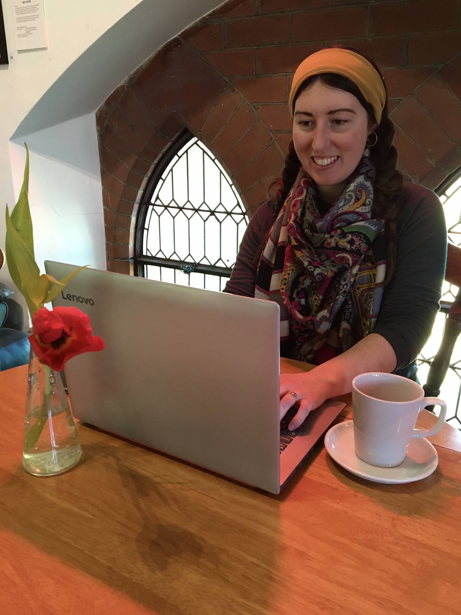 A visitor working on a Lenovo laptop beside a tall Gothic stained-glass arched window, sunlight on the table, tulip in a small vase, a cup of tea beside her.