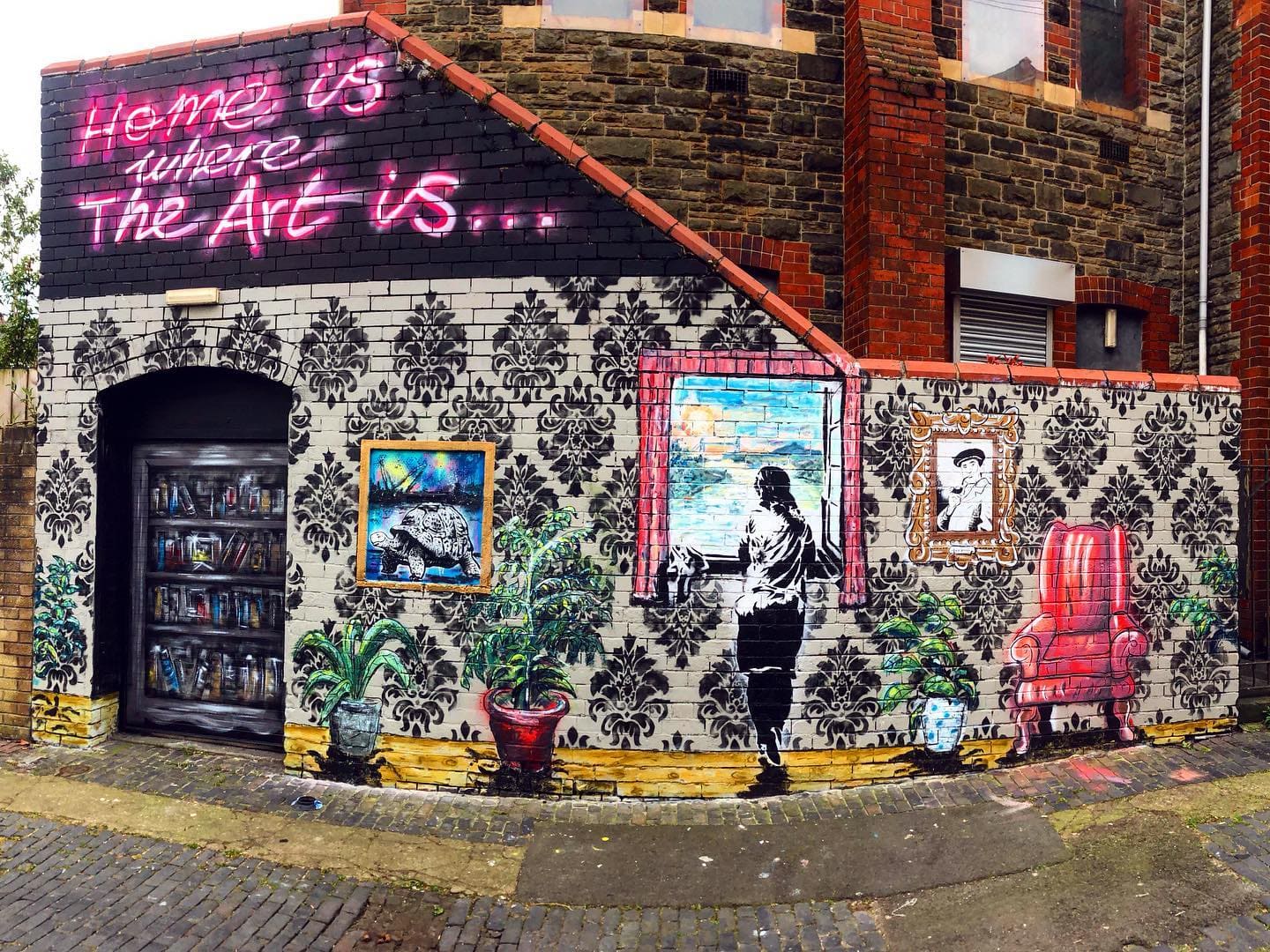 Ruperra Lane mural project beside Barnabas Arts House. "Home is Where the Art Is" hand-painted in pink across an old brick wall, with framed paintings, a figure and an armchair painted into the mural beneath.