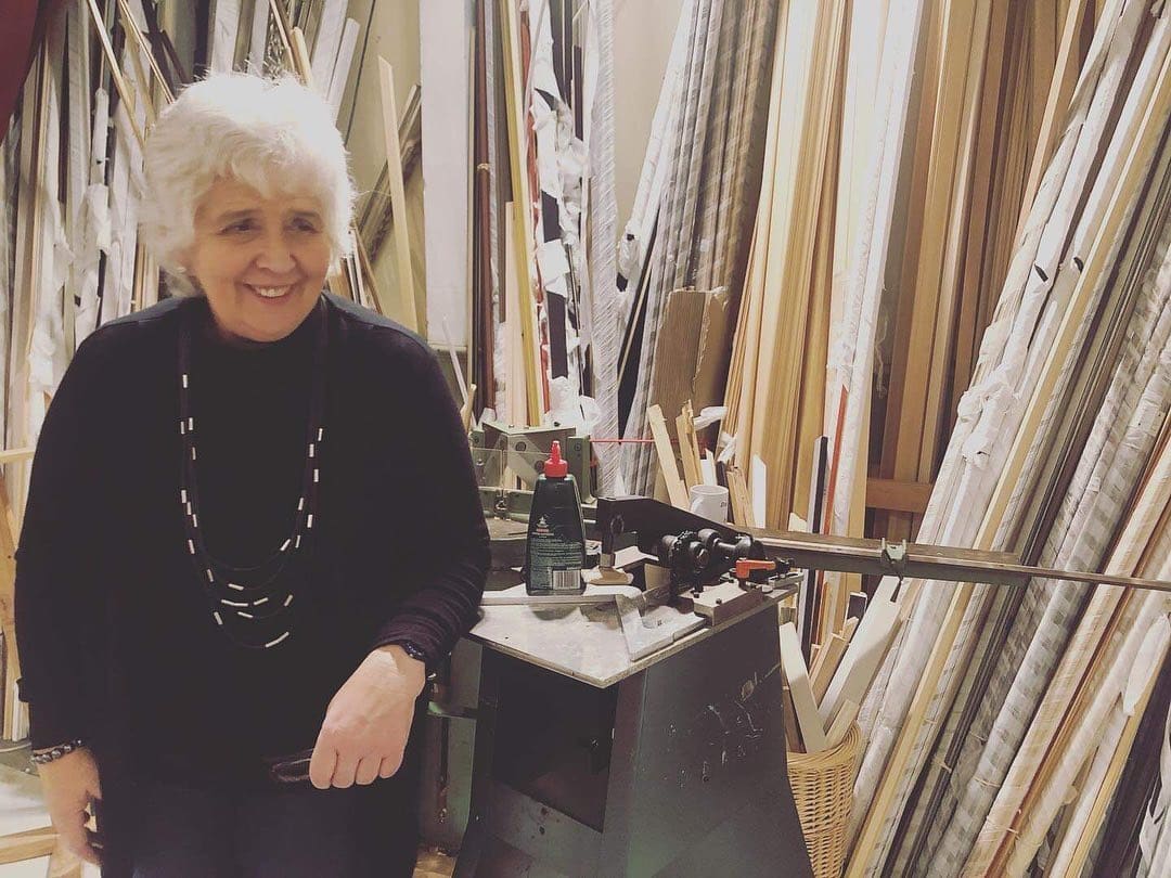 Janet Martin, founder of Barnabas Arts House, smiling beside the mounting machine in the Gwent Picture Framing workshop, surrounded floor-to-ceiling by stacks of mouldings in walnut, oak, painted white and gilt.
