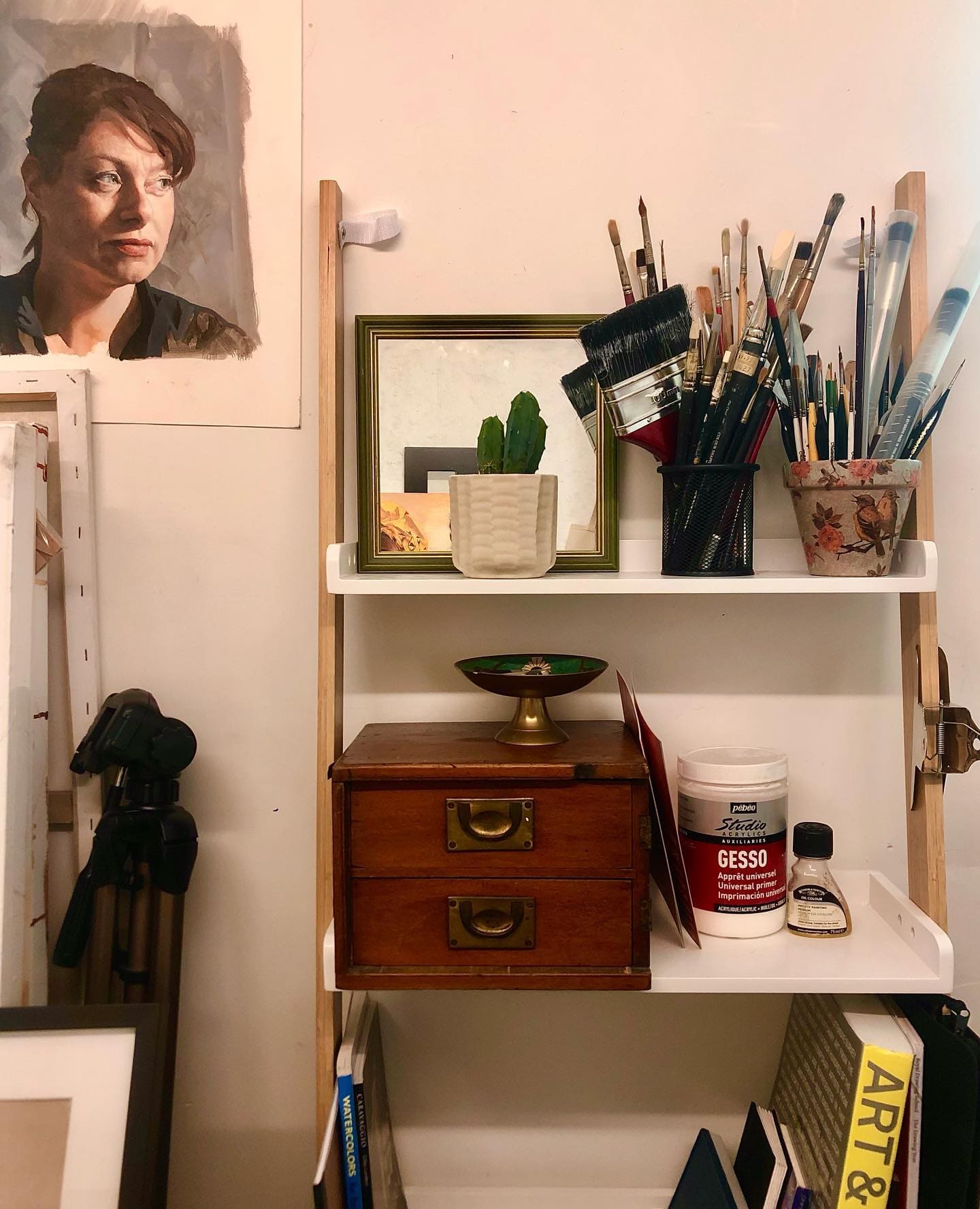 A resident artist's shelf inside Barnabas Arts House. Paintbrushes in jars, a Pebeo gesso tin, a watercolours book, an unfinished portrait pinned above an antique wooden chest of drawers.